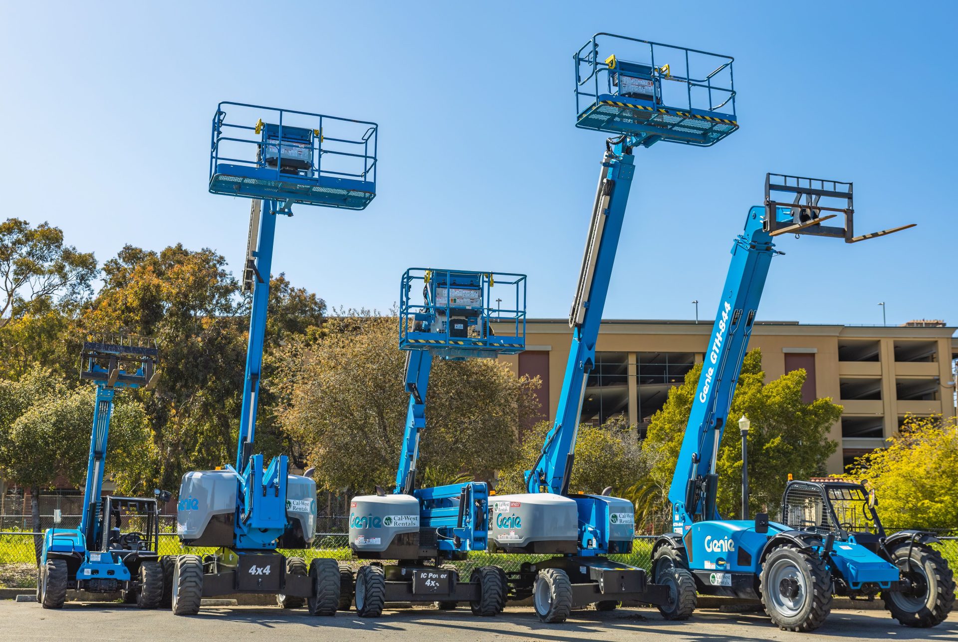 Scène extérieure avec plusieurs nacelles élévatrices bleues et des engins de chantier dans une zone industrielle ensoleillée.